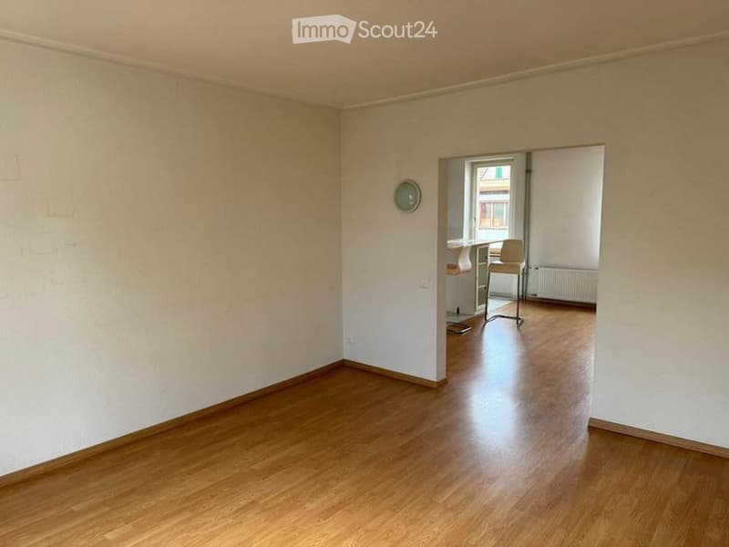 Empty room with wooden flooring and white walls, featuring a doorway leading to another room with a small table and chairs.