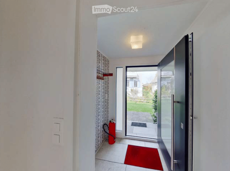 Modern hallway with white walls, a red doormat, and a glass door leading to a garden area. A red fire extinguisher is visible on the wall.