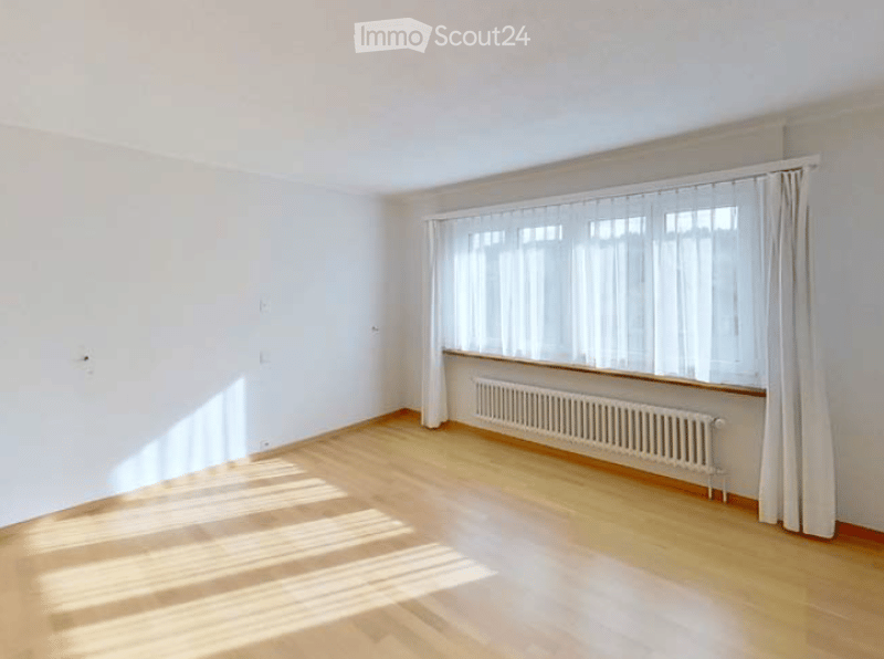 Completely empty room with white walls, wooden floor, large window with white curtains, and radiator below the window.