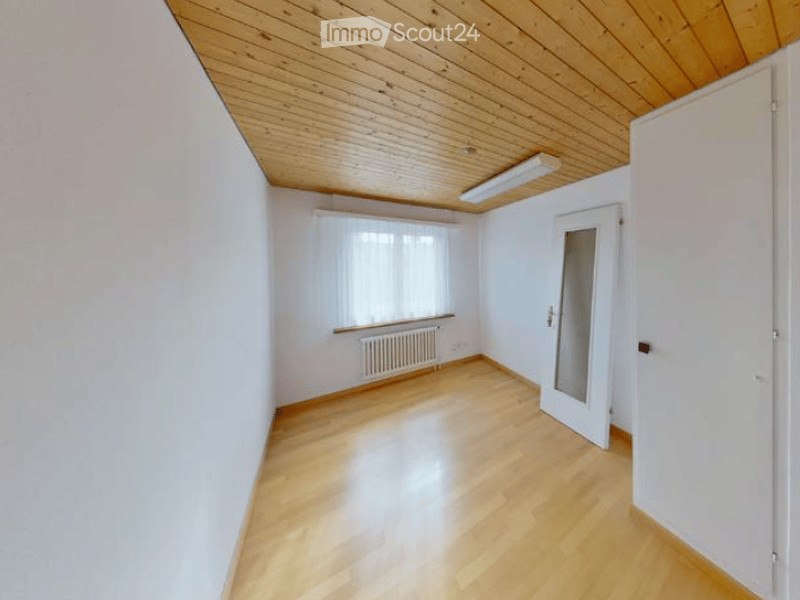 Empty room with wooden flooring, white walls, and a wooden ceiling. Features include a window with blinds, a radiator, and a white door.