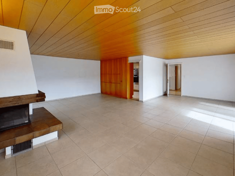 Empty room with white walls, tiled floor, and a fireplace on the left side. Wooden cabinetry is visible in the background, and the ceiling has a unique wooden design.