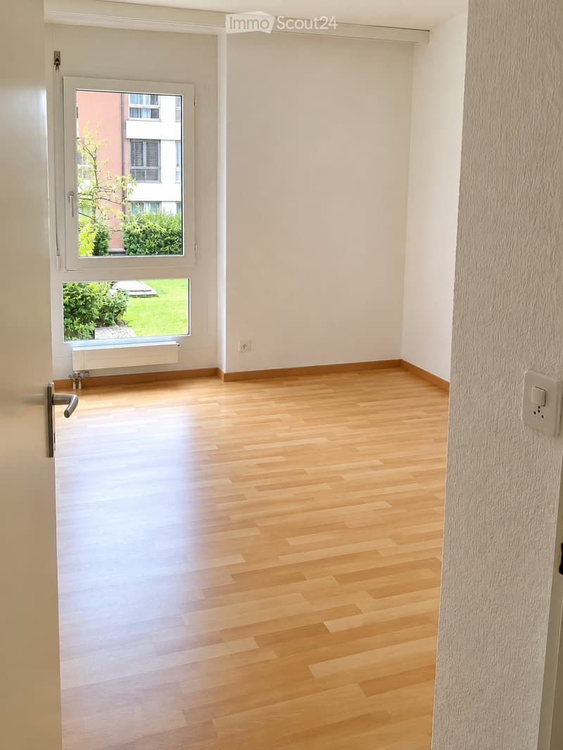 Empty room with light wooden flooring, white walls, and a large window offering a view of greenery and a neighboring building.