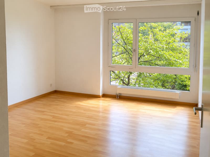 Empty room with light wooden flooring, white walls, and large windows showing greenery outside.