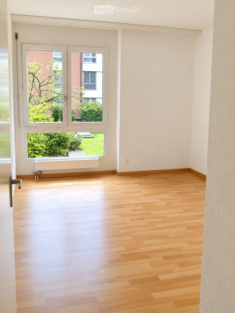 Empty room with light wooden flooring, white walls, large windows with a view of greenery, and a radiator below one window.