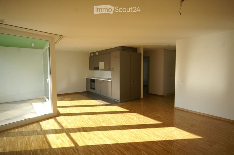 Empty kitchen with wooden flooring, grey cabinets, and a large window letting in natural light.