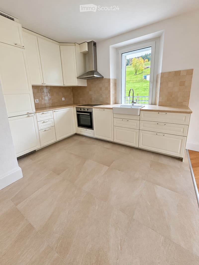 Modern kitchen with white cabinets, beige tiles, and a large window overlooking a green landscape. Features include a sink, oven, and range hood.