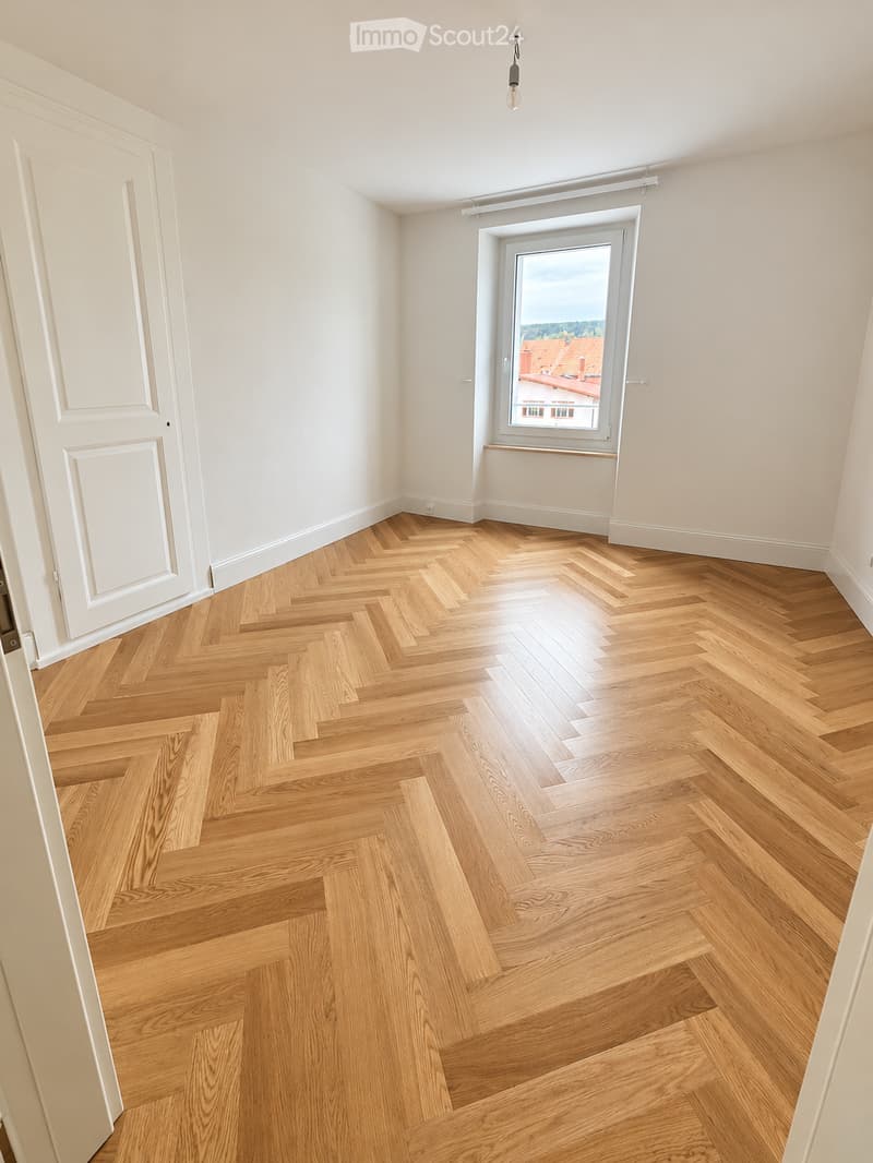 Empty room with light wooden herringbone flooring, white walls, a white door, and a large window with a view of rooftops and greenery.