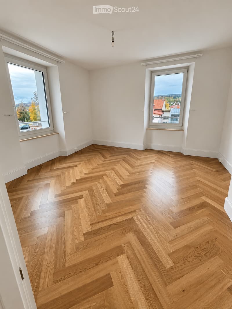 Empty room with herringbone-patterned wooden flooring, white walls, and large windows offering a view of the outside. No furniture or furnishings are present.