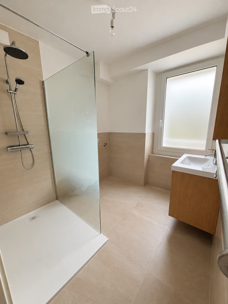 Modern bathroom with a glass-enclosed shower, a single sink with a wooden cabinet, and a large window providing natural light.