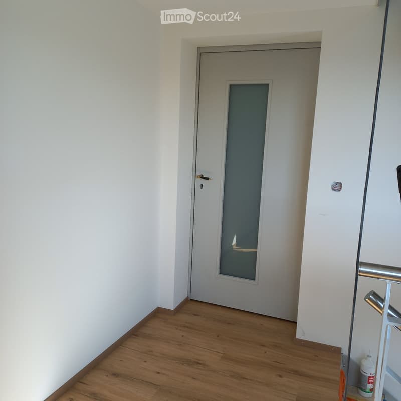Empty hallway with wooden flooring, white walls, and a closed door with a small window. A cleaning spray bottle is visible on the floor near the door frame.