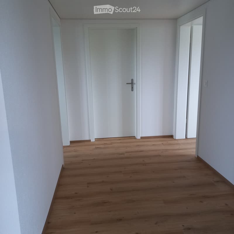 Empty hallway with light wooden flooring and white walls, featuring a closed door and an open doorway leading to another space.
