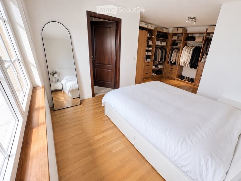 Modern bedroom with a large bed, built-in wardrobe, full-length mirror, nightstand, and lamp. Wooden flooring and large windows provide natural light.