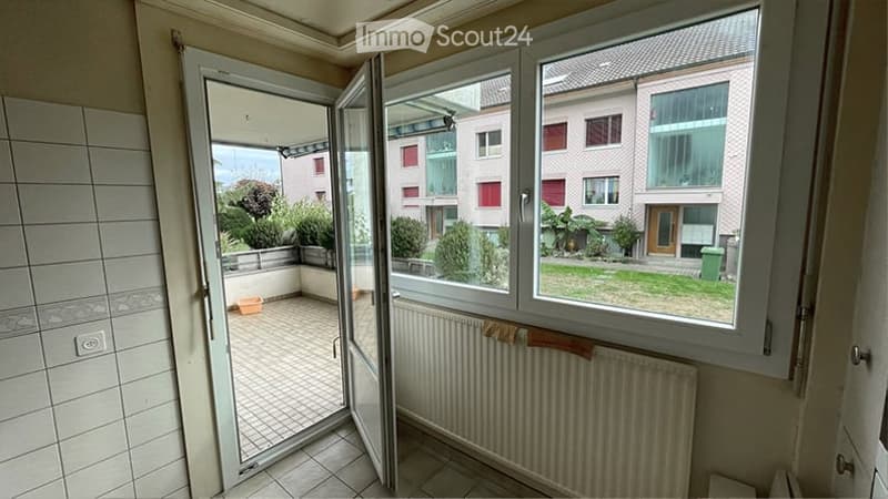 Balcony with sliding glass doors, tiled floor, radiator, and view of a garden and neighboring buildings.