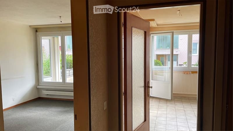 Empty room with carpeted floor, large window with white frame, radiator below window, and an open door leading to another room with tiled floor and window view.