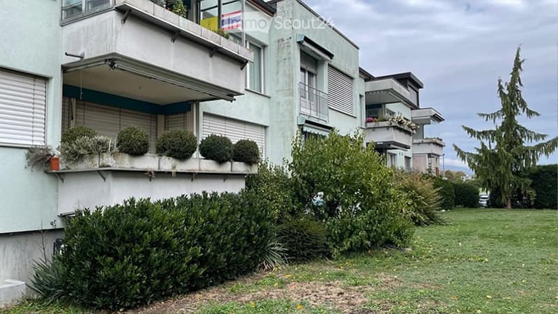 Modern multi-story apartment building with balconies, greenery, and a well-maintained lawn in front.
