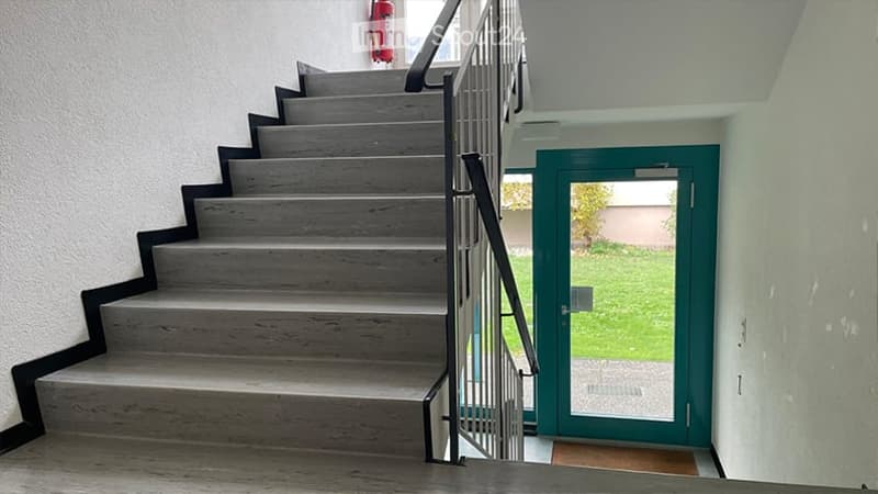 A modern staircase with grey wooden steps and a black metal railing, leading up to a green double door that opens to a garden area.
