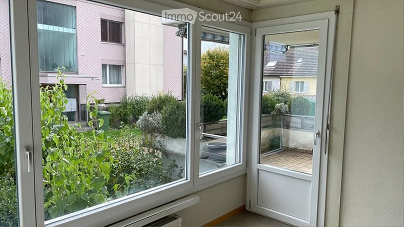 Balcony with glass doors, overlooking a garden area with greenery and neighboring buildings.