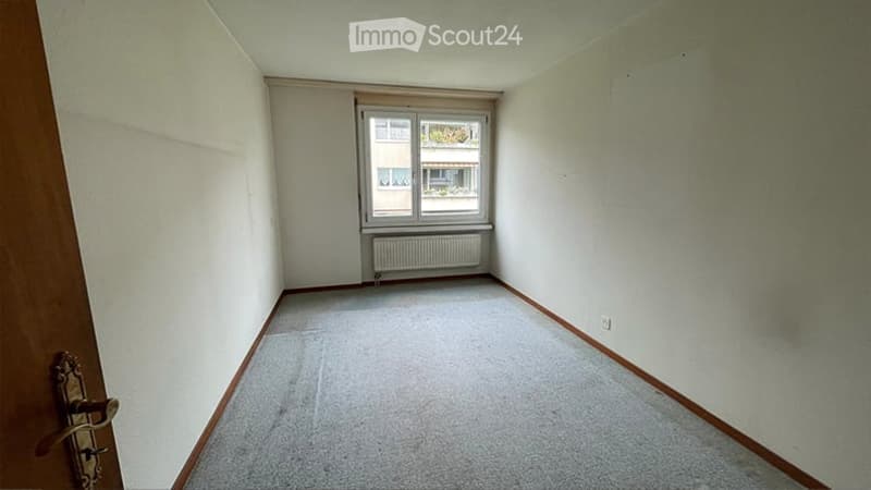Empty room with light-colored walls, carpeted floor, and a single window with a radiator below it. The room has a wooden door frame on the left side.