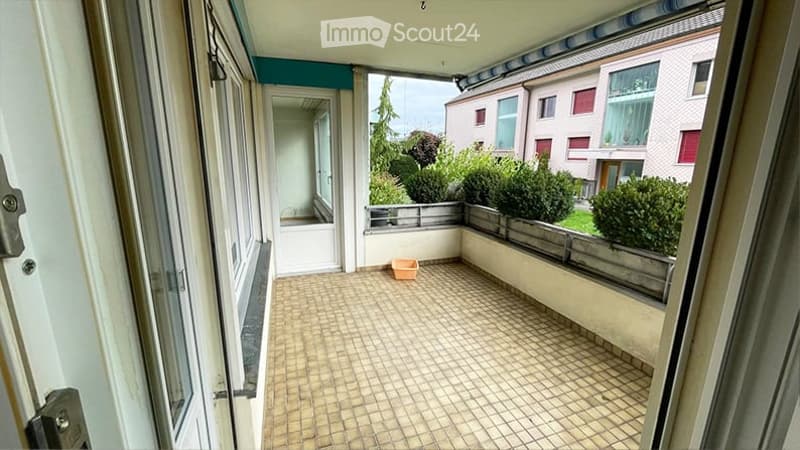 Balcony with tiled floor, metal railing, small orange container, view of neighboring buildings and greenery.
