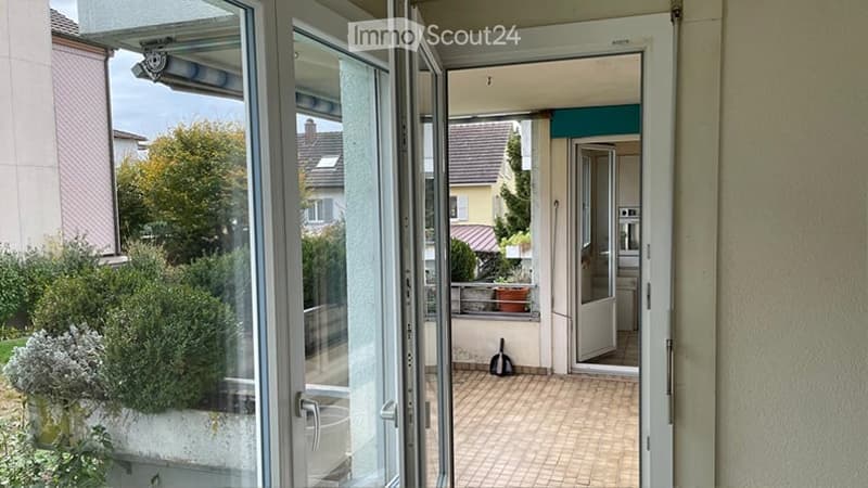 Balcony with tiled floor, glass doors leading to a hallway, potted plants, and a view of neighboring buildings and greenery.