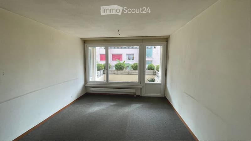 Empty room with grey carpet, white walls, large glass door leading to a balcony, radiator, and a view of greenery outside.