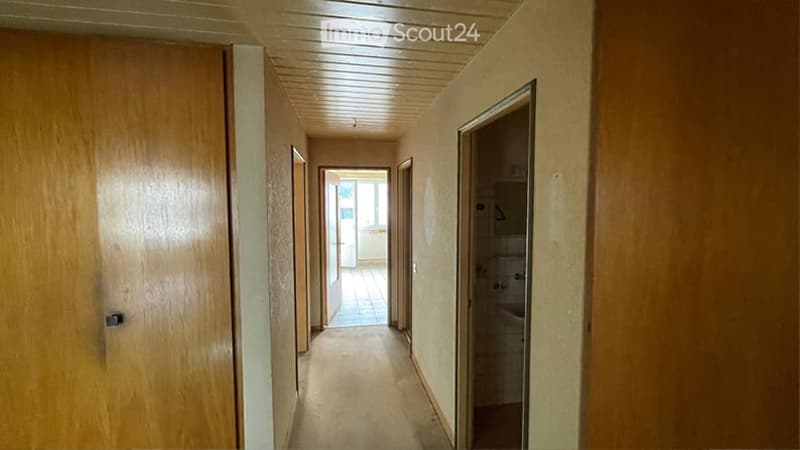 A narrow hallway with wooden doors on both sides, leading to a partially visible bathroom and another room. The ceiling is paneled, and the floor is tiled.