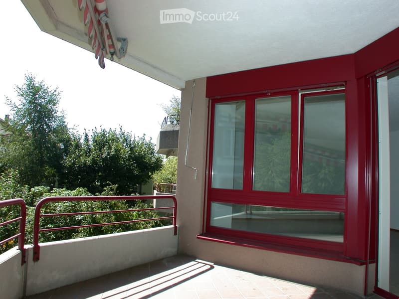 Balcony with red-framed windows, metal railing, and outdoor umbrella hanging from the ceiling. The view shows greenery and a neighboring building.