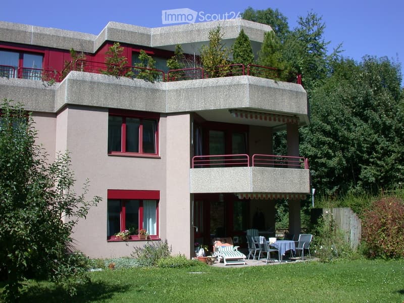 Two-story house with red-framed windows, balconies, and a well-maintained garden with outdoor furniture.