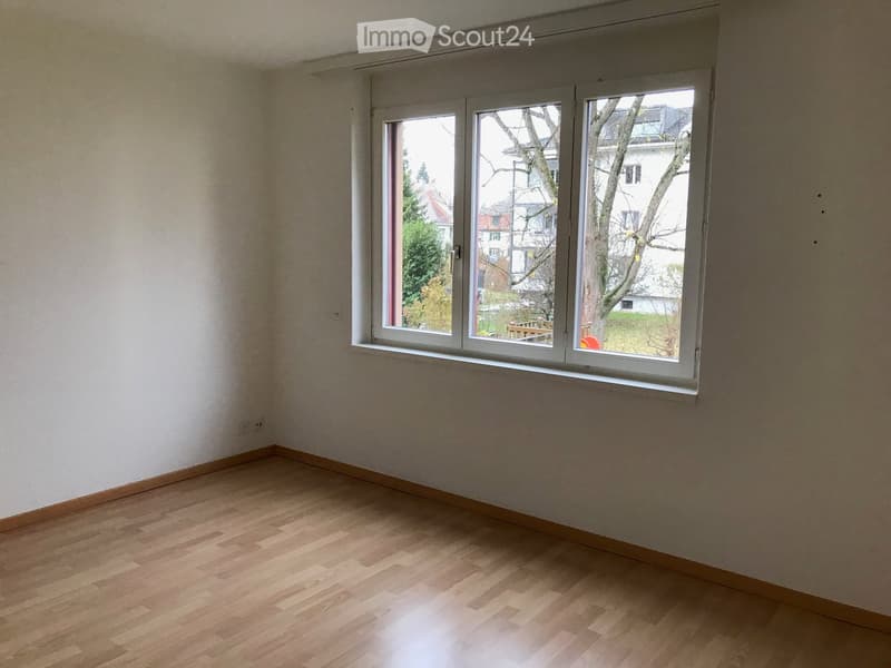 Empty room with wooden flooring and large windows showing a view of the outside area.