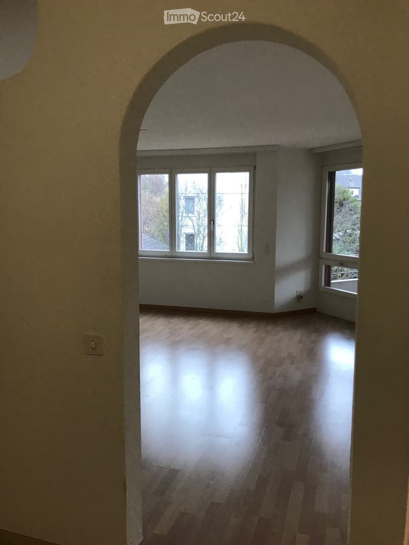 Empty room with wooden flooring, large windows, and an arched doorway leading to another space.
