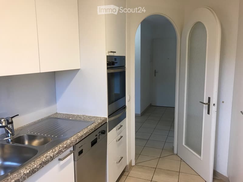 Modern kitchen with white cabinets, granite countertop, stainless steel sink, and built-in oven and dishwasher. An arched doorway leads to a tiled hallway.