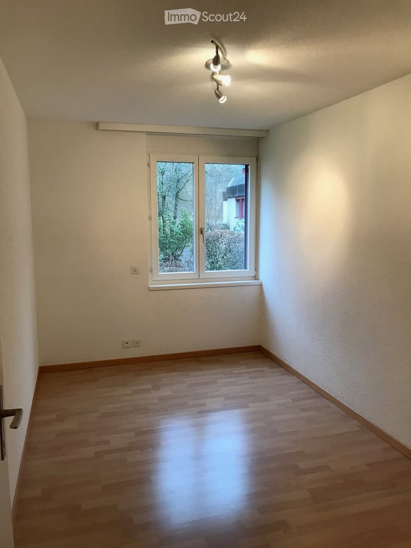 Empty room with wooden flooring, white walls, and a large window with blinds. The room has a modern ceiling light fixture and electrical outlets on the walls.