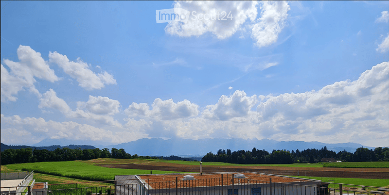 A scenic view of a rural landscape with green fields, scattered trees, and distant mountains under a partly cloudy sky.