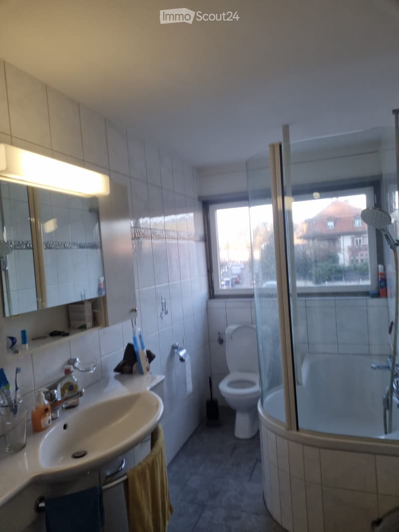 Bathroom: toilet, glass shower, large window, white tiles, mirror with light, sink, yellow towel, bathroom accessories.