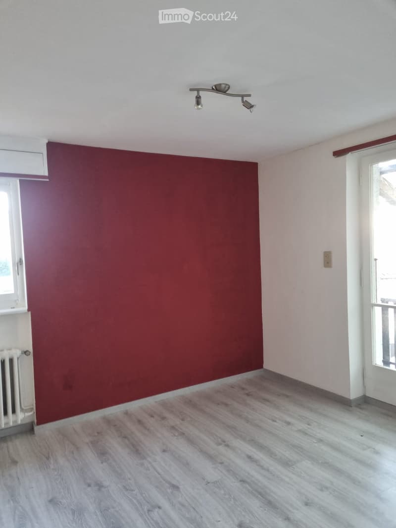 An empty room with a red wall, a white floor, a door on the left, a window on the wall, a ceiling with lights, a white radiator, and a balcony visible through a door.