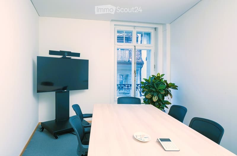 Conference room with a large screen on a rolling stand, a long wooden table surrounded by chairs, a potted plant, and a calculator on the table.