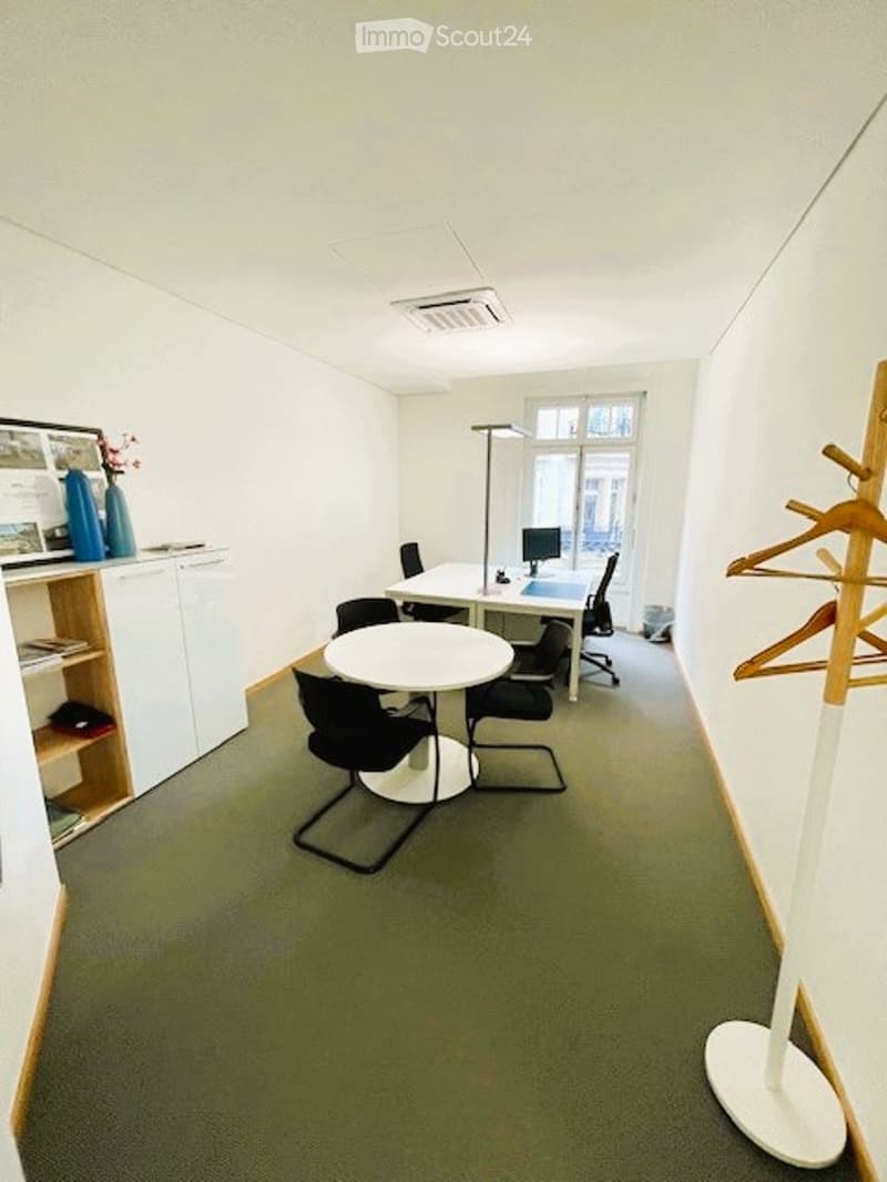 A modern, well-lit office space with white walls, green carpeting, and large windows. It features a round table with four chairs, a desk with a computer screen, and a cabinet with a vase and books.