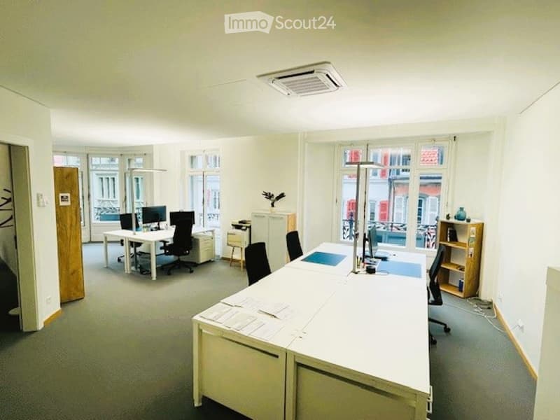 Bright office space with white desks, black chairs, computer monitors, and shelves. Natural light from large windows.