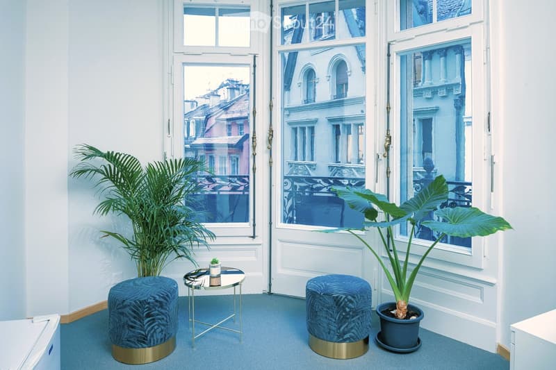 A small, modern room with blue carpeting, two blue velvet stools, a small round table, and two potted plants near large windows with a view of the city.