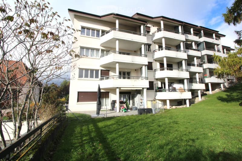 A modern, multi-story residential building with white exterior walls, multiple balconies, and large windows. The building is surrounded by a well-maintained lawn and trees.