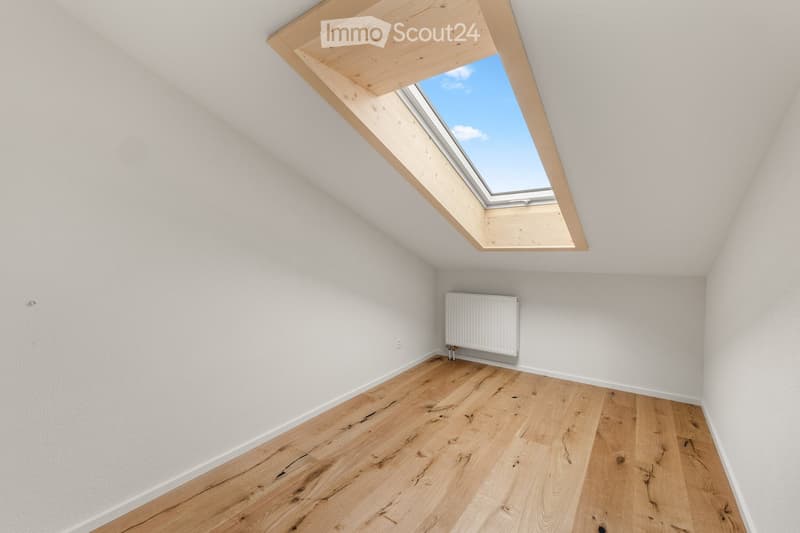 Empty attic room with wooden flooring, white walls, and a skylight window at the top.