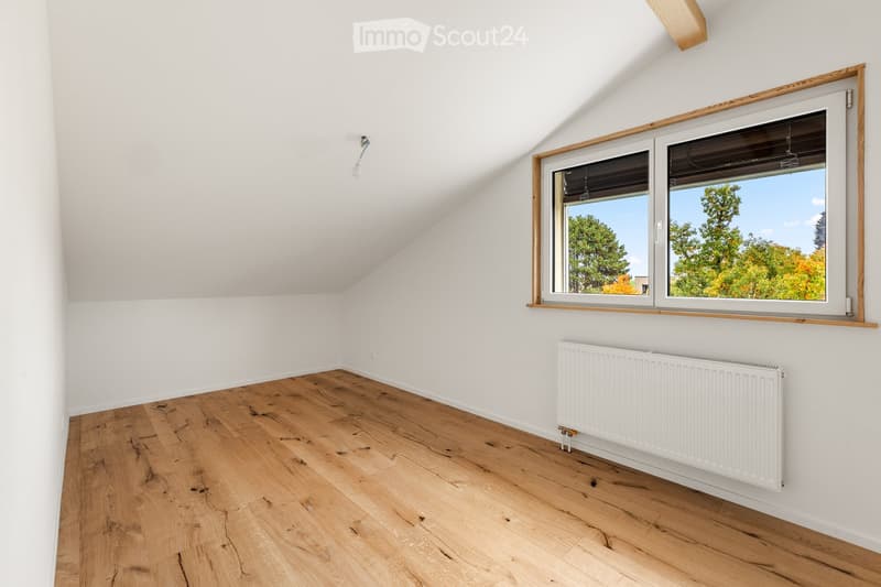 An empty attic room with wooden flooring, white walls, and a large window offering a view of trees and sky. A white radiator is mounted on the wall below the window.