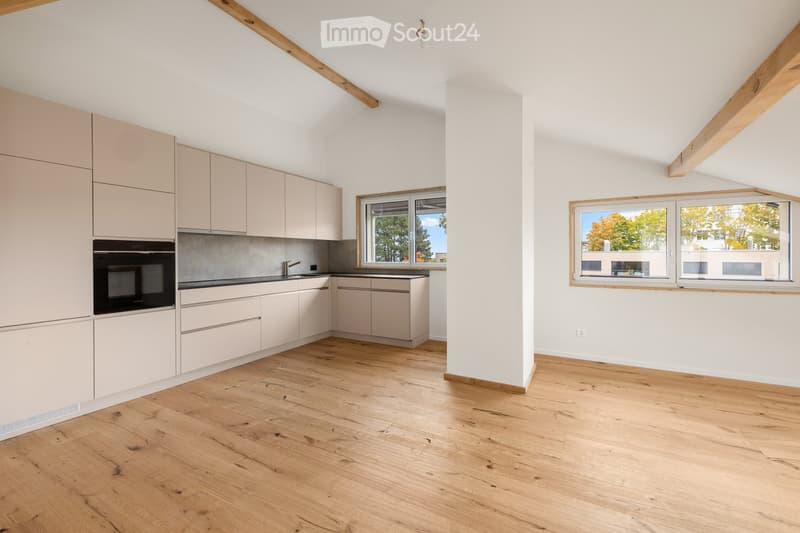 Bright, open room with wooden flooring, white walls, and large windows offering natural light. The space includes a kitchen area with light-colored cabinets and a built-in oven, but no furniture or personal items are present.
