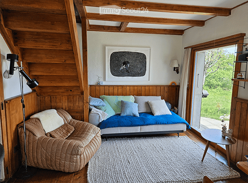 Cozy living room with wooden beams, a blue sofa with multiple pillows, a beige armchair, a small wooden table, and a floor lamp. The room features exposed wooden beams and a large window with a view of greenery.