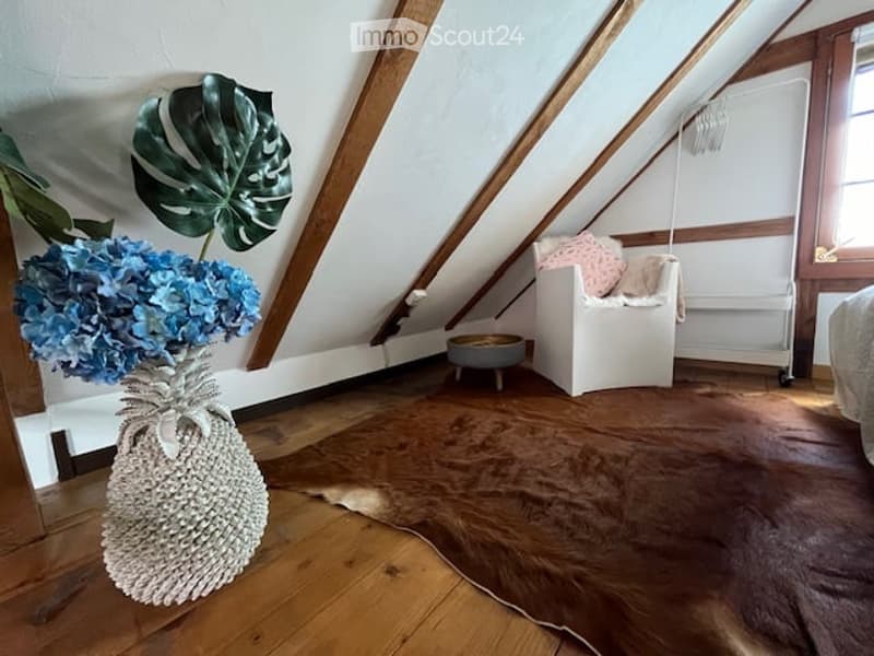 A room with wooden floors, wooden beams, a rug, a white chair, a round table, a plant, and blue flowers in a vase.