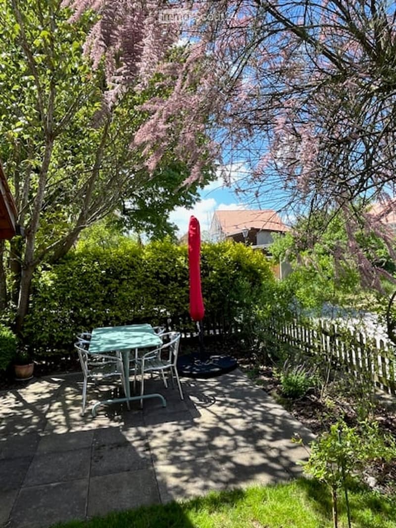 Outdoor area with table and chairs, surrounded by greenery, red umbrella, and fence, with a view of a house and distant buildings