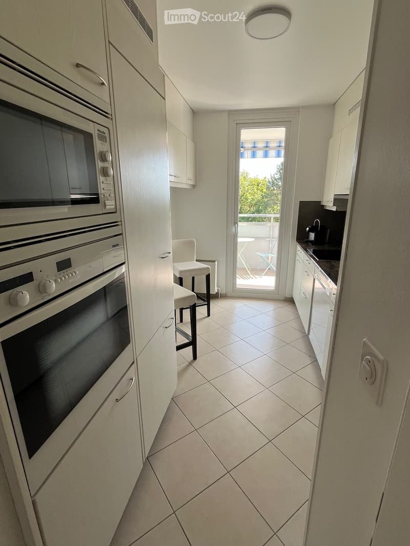 Modern kitchen with white cabinets, tiled floor, built-in oven, sink, and a small white stool near the balcony door.
