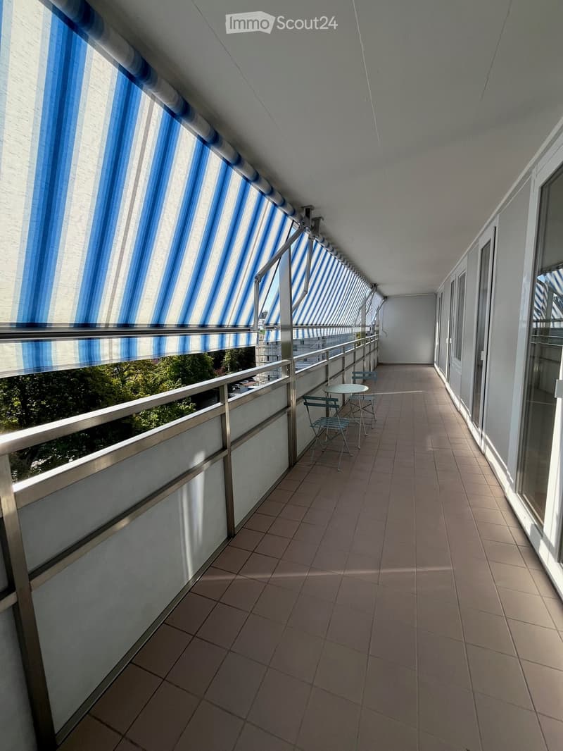 Balcony with tiled floor, metal railing, and a small table with chairs. Blue and white striped awnings cover the balcony.
