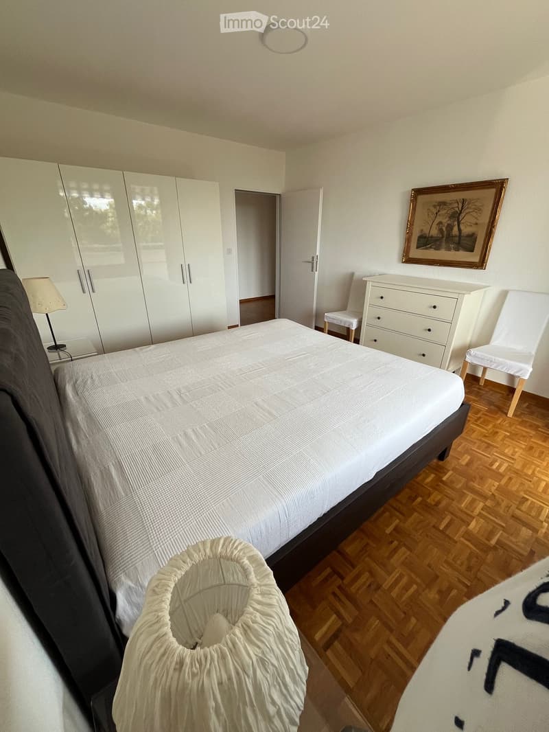 Modern bedroom with a large bed, white wardrobe, a small table with a lamp, a chair, and a framed picture on the wall.