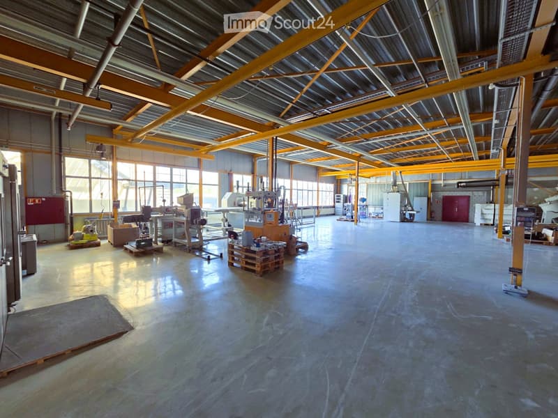 Large industrial-style warehouse with a high ceiling, metal beams, and large windows allowing natural light. The floor is concrete, and there are various industrial equipment and storage items scattered throughout.